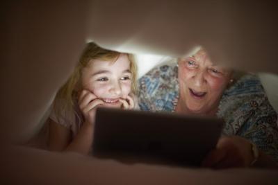 A child and her grandmother under covers, their smiling faces lit up by a screen.